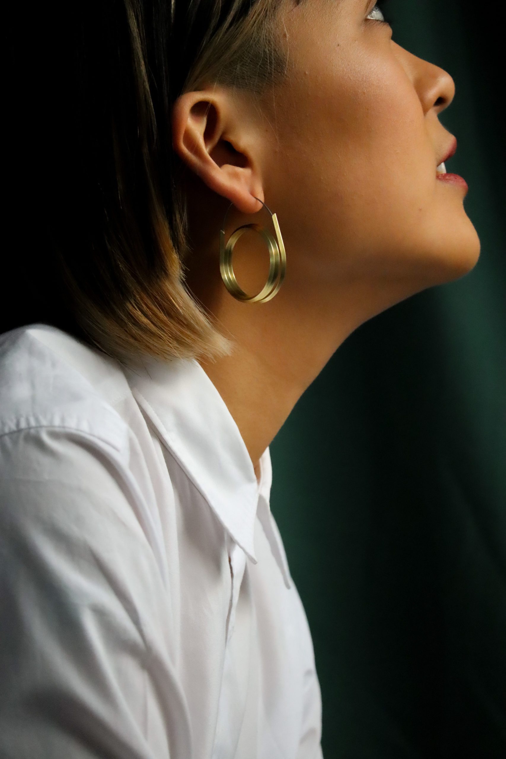 Photo of a side on view of a young woman from the shoulders up wearing a white collared shirt, double brass hoops, looking up, on a green background
